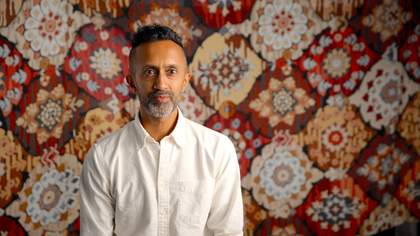 Artist Hetain Patel sits facing the camera with a colorful patterned carpet behind him