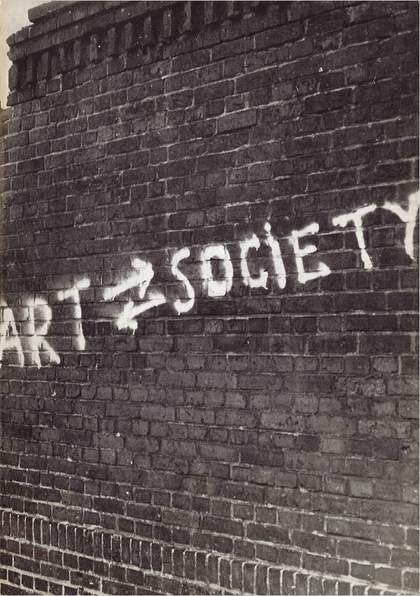 Black-and-white photograph of a brick wall with white graffiti on it reading ART SOCIETY with two arrows between the two words, the upper pointing right (towards SOCIETY), the lower pointing left (towards ART) 