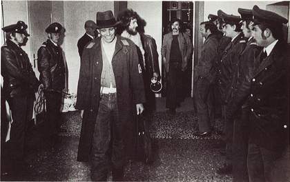 Black-and-white photograph of Joseph Beuys in hat and coat, grinning, walking towards the camera. The room is lined on both sides with policemen.