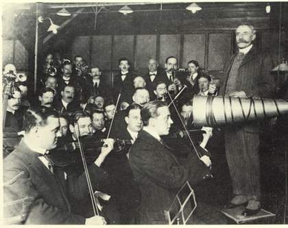 A black and white photograph of a figure standing in front of an orchestra and next to a large, cone-shaped horn