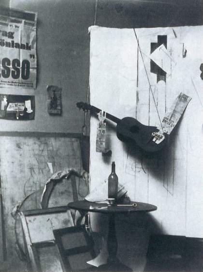 A large white sheet with abstract lines drawn onto it, with a guitar and newspaper hanging down and resting on its surface and a table and wine bottle positioned in front of it