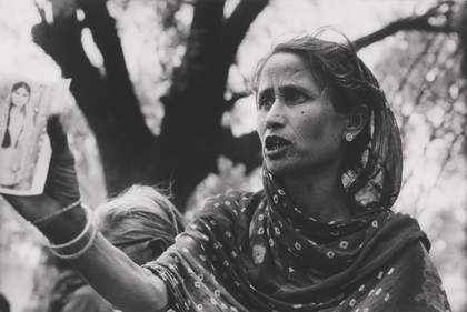 A figure stands in an outdoor setting with her mouth open as if speaking, and holds a photograph of a person with long hair.