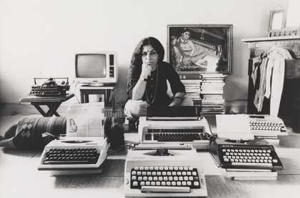 A figure sits cross-legged on the floor in an indoor setting, their chin resting on their hand, surrounded by typewriters, a television, a painting of a seated figure and a chest of drawers with cloth spilling out of it.