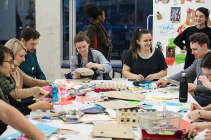 A group of people sit around a table making objects using the craft materials that are in front of them.