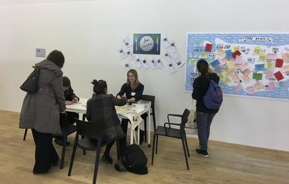 A small group of people sit writing and talking at a table, while another person looks at a large wall map with coloured pieces of paper pinned to it.