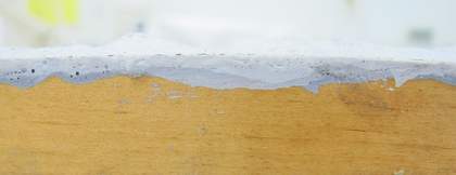 A horizontal view of the edge of a piece of wood with a thin layer of darker grey paint on it and a layer of lighter grey paint over the top.