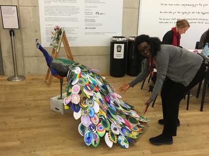 A person faces the camera and presents a large sculpture of a peacock with an array of brightly coloured feathers.