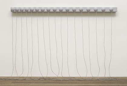 A grey sculpture consisting of a narrow, horizontal panel with seventeen hemispheres on its face, fixed to the wall around two metres above the floor, with long ropes from the centre of each hemisphere that hang down and rest in coils on the floor