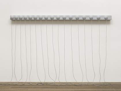 A grey sculpture consisting of a narrow, horizontal panel with seventeen hemispheres on its face, fixed to the wall around two metres above the floor, with long ropes from the centre of each hemisphere that hang down and rest in coils on the floor