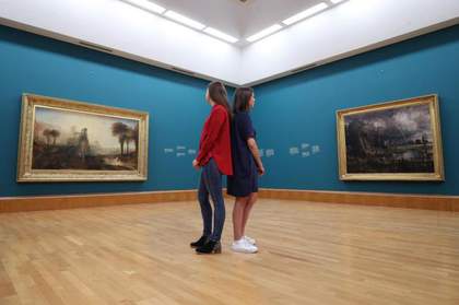 Photograph of the two works in straight, brightly gilded frames on either side of a corner in a gallery with blue walls.