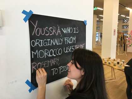 A person writes on a blackboard with coloured chalk. The text reads ‘Youssra, who is originally from Morocco, uses thyme rosemary…’