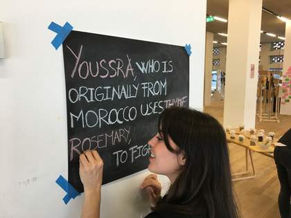 A person writes on a blackboard with coloured chalk. The text reads ‘Youssra, who is originally from Morocco, uses thyme rosemary…’
