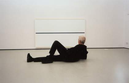 A photograph of Uglow reclining in a gallery, looking at a canvas with a single horizontal line across its middle.