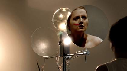 Film still shot from behind as a woman looked in one of three round mirrors. She has a wet red line from her nose to her cheek which could be lipstick, paint or blood.