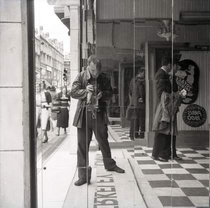 Black and white photograph of Nigel Henderson taking a picture of himself in a mirrored wall by a busy street