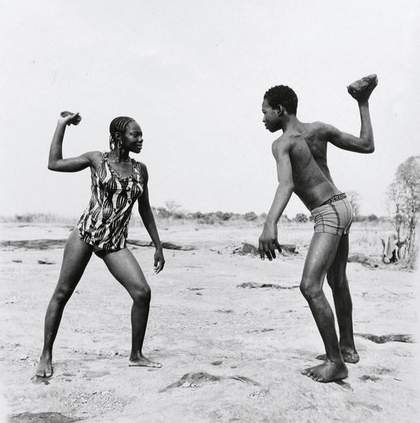 Malick Sadibe Friends Fighting with Stones 1976 black and white photograph of a male and female figure set in the African countryside each holding a stone above their heads and facing one another 