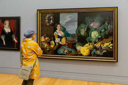 A person looks at a large painting featuring a woman and many vegetables