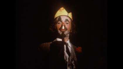 a man with clown-like make up on stands with a crown