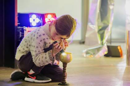 A child kneeling on the ground holding a torch and an eyeglass over a gold goblet, exploring the object with light